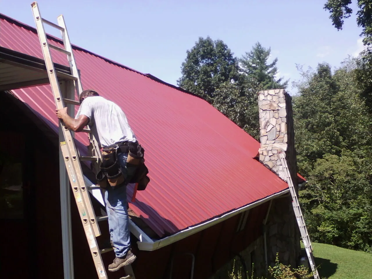 Roofer on a ladder beside a red metal roof and stone chimney — Soffit & Fascia Repair by Roofing Brothers in Swan